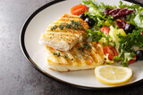 Diet food grilled fish cod with fresh salad of tomatoes, olives, arugula, lettuce mix and lemon close-up in a plate on the table. Horizontal