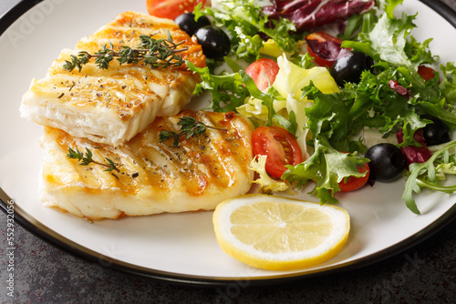 Delicious healthy grilled cod fish with fresh salad of tomatoes, olives, lettuce mix and lemon close-up in a plate on the table. Horizontal