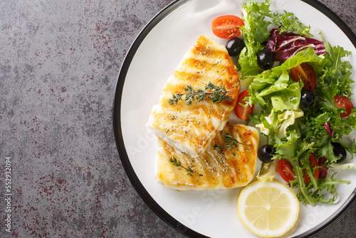 Delicious healthy grilled cod fish with fresh salad of tomatoes, olives, lettuce mix and lemon close-up in a plate on the table. Horizontal top view from above