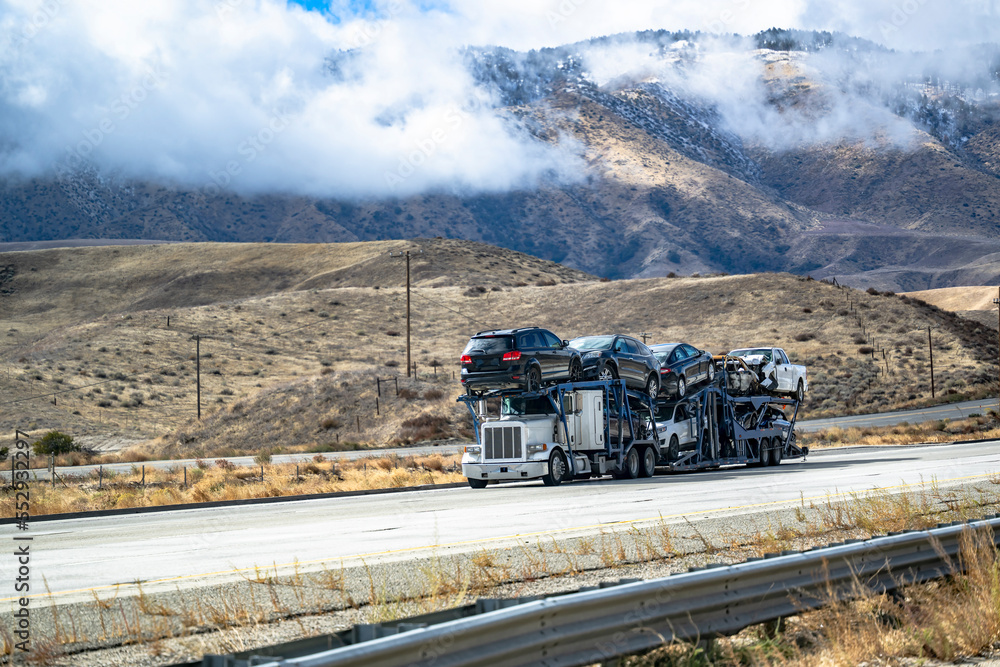 Industrial classic big rig white car hauler semi truck transporting heavy cars on the modular semi trailer driving on the divided highway road with snow mountains hidden in the clouds