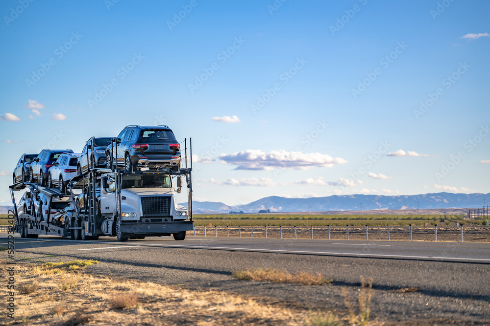 Industrial grade low cab big rig semi truck transporting cars on the hydraulic semi trailer driving on the flat California plato
