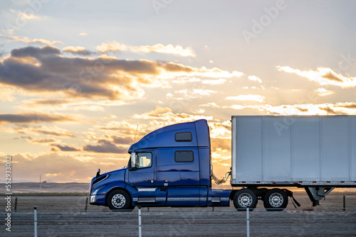 Profile of big rig blue semi truck tractor with dry van semi trailer driving on the flat highway road along the fields at twilight