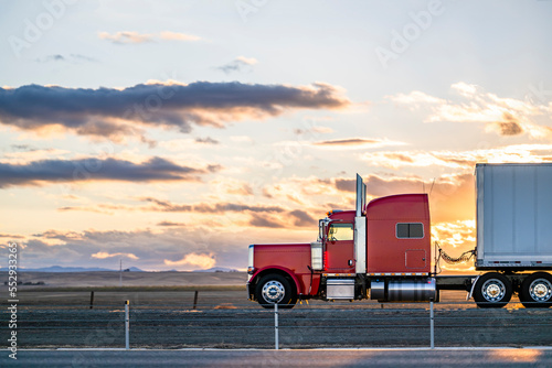 Side view of red big rig classic semi truck transporting cargo in dry van semi trailer moving on the road at sunset