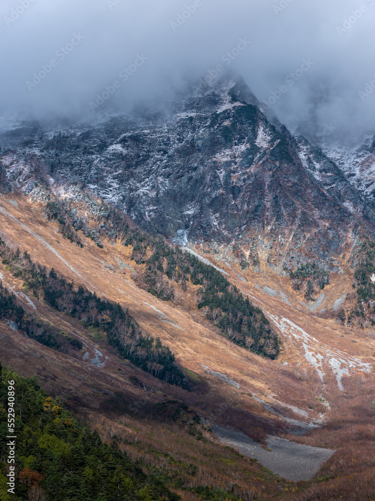 晩秋のアルプス山脈に初雪の山肌が目を引く、山頂は雲に隠れ、紅葉か残る渓谷にスポットライトが当たり黄色く輝いている。