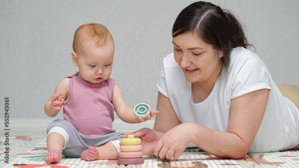 Baby girl enjoys playing pyramid toy with mother on floor. Brown-haired woman teaches excited daughter to put toys in right order and praises child work Baby girl enjoys playing pyramid toy with mother on floor. Brown-haired woman teaches excited daughter to put toys in right order and praises child work