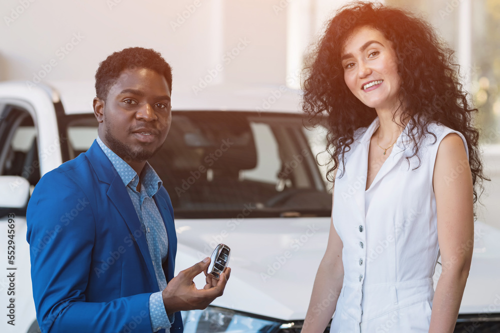 Obraz premium African American man dealer gives black-haired female customer key to purchased car. Businesswoman looks with satisfied expression and enjoys new purchase, sunlight