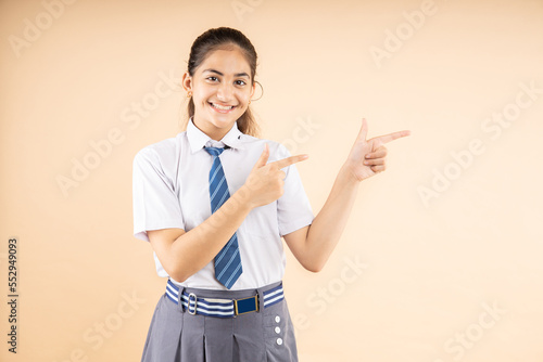 Happy Indian student schoolgirl wearing school uniform pointing at blank space to put advertisement isolated over beige background, Studio shot, closeup, Education concept.