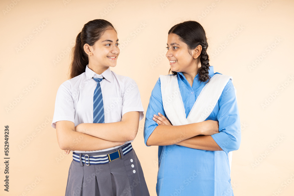 Happy Rural and Modern Indian student schoolgirls wearing school uniform standing together cross arms looking at each other isolated over beige background, Closeup, Studio shot, Education concept. Happy Rural and Modern Indian student schoolgirls wearing school uniform standing together cross arms looking at each other isolated over beige background, Closeup, Studio shot, Education concept.