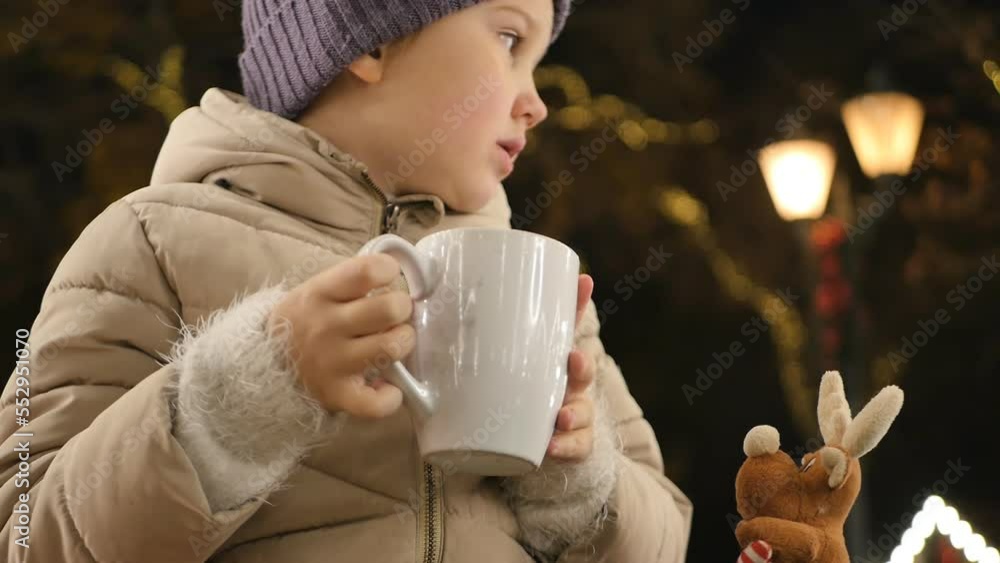 A cute and kind girl dressed in a winter jacket and a hat at night in the park drinks a hot drink holding a mug and laughs exhaling steam from her mouth into the cold winter air A cute and kind girl dressed in a winter jacket and a hat at night in the park drinks a hot drink holding a mug and laughs exhaling steam from her mouth into the cold winter air