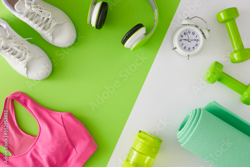 Fitness concept. Flat lay photo of dumbbells sports shoes bottle of water exercise mat pink sports top headphones and alarm clock on white and green background with copy space in the middle.