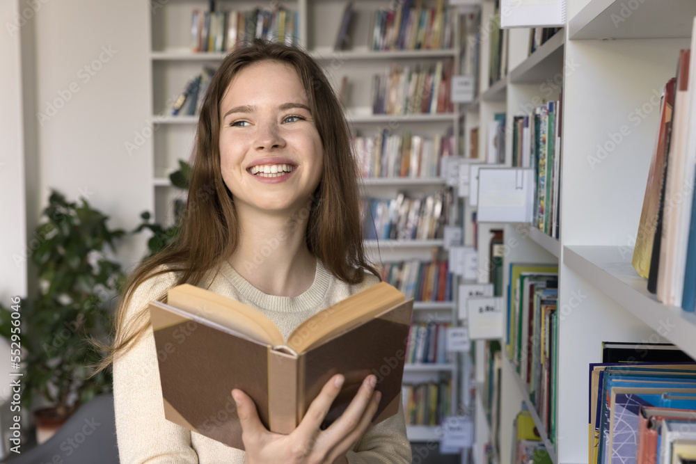 Happy pretty student girl excited with reading, holding open book, standing at bookshelf in college, university, school library, enjoying studying, education, getting knowledge Happy pretty student girl excited with reading, holding open book, standing at bookshelf in college, university, school library, enjoying studying, education, getting knowledge