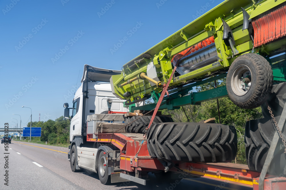 Heavy industrial truck with low side on low-frame platform transports disassembled big green combine harvester along ordinary highway on summer day.