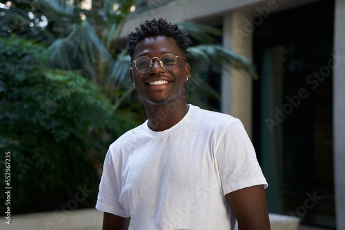 Portrait of black man very happy, smiling and looking at camera. An African American student sincerely smiles while standing outdoors. The guy is wearing a white t-shirt