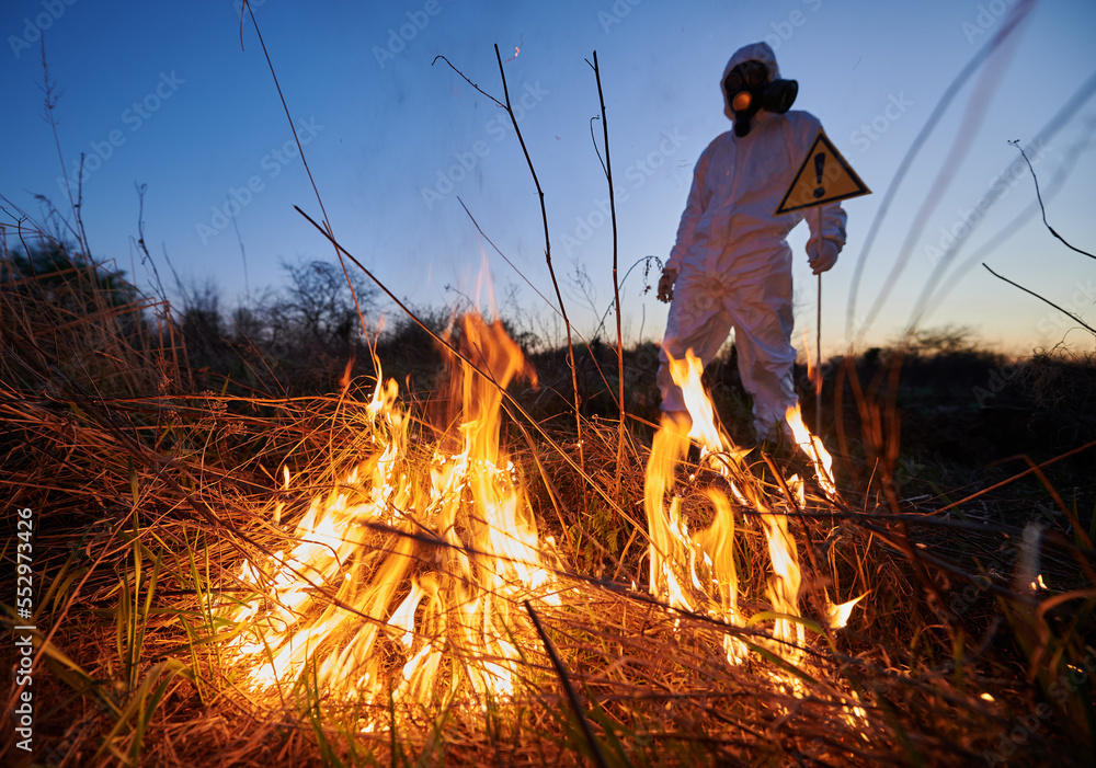 Fireman ecologist working in field with wildfire at night. Man in protective suit and gas mask near burning grass with smoke, holding warning sign with exclamation mark. Natural disaster concept. Fireman ecologist working in field with wildfire at night. Man in protective suit and gas mask near burning grass with smoke, holding warning sign with exclamation mark. Natural disaster concept.
