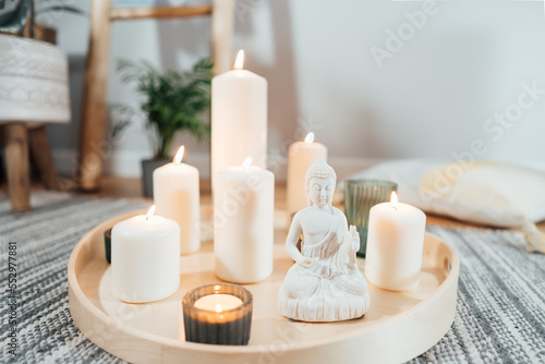 Wooden tray with burning candles and white Buddha statuette on the floor of modern Scandi interior. Zen Composition for meditation, yoga practice, relaxation. Balance and calm energy flow indoor.