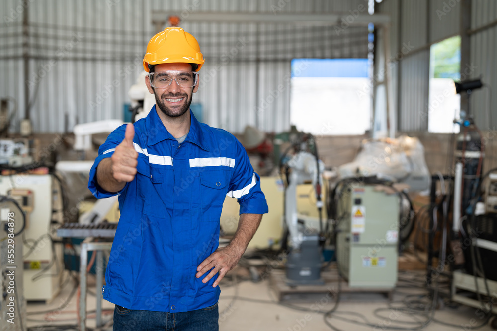 Portrait of male engineer in uniform smiling and showing thumbs up at industrial factory. Technician man standing wearing yellow helmet safety in manufacturing workshop. Portrait of male engineer in uniform smiling and showing thumbs up at industrial factory. Technician man standing wearing yellow helmet safety in manufacturing workshop.
