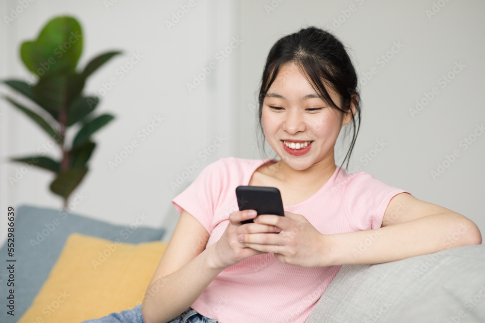 Excited korean lady sitting on sofa and using smartphone, chatting with friends online, checking social media Excited korean lady sitting on sofa and using smartphone, chatting with friends online, checking social media