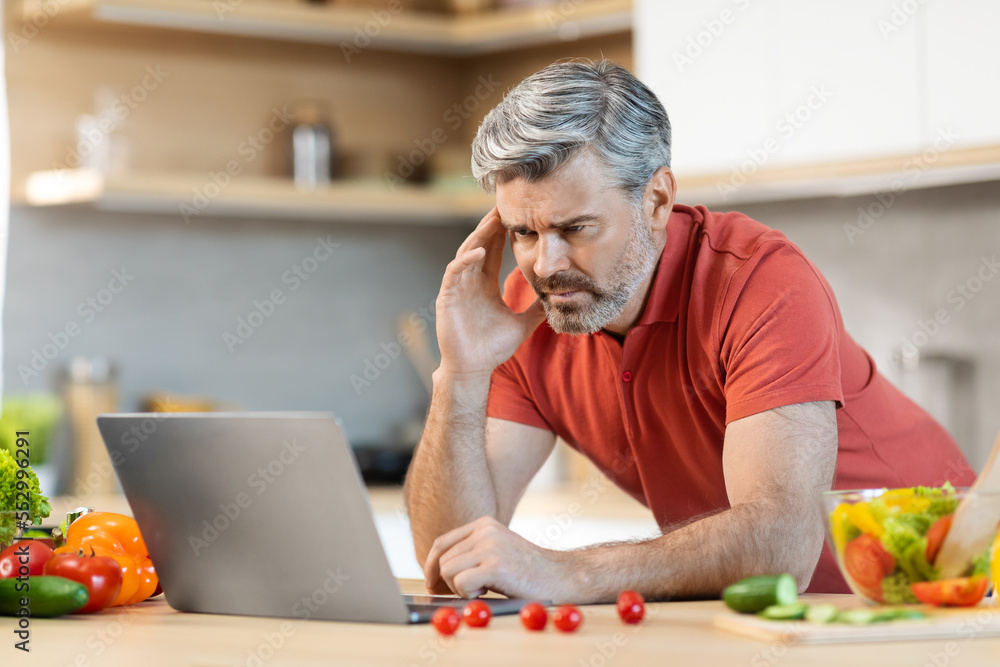 Stressed middle aged man looking at computer screen while cooking Stressed middle aged man looking at computer screen while cooking