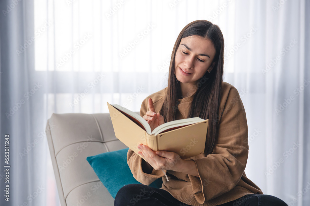 A young woman is reading a book while sitting on an armchair by the window. A young woman is reading a book while sitting on an armchair by the window.