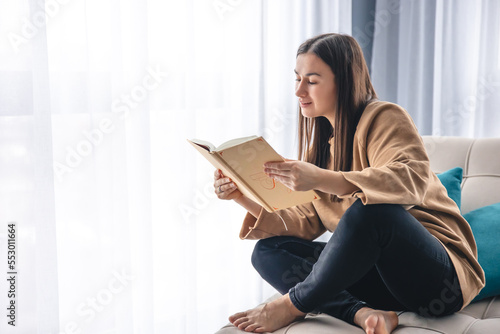 A young woman is reading a book while sitting on an armchair by the window.
