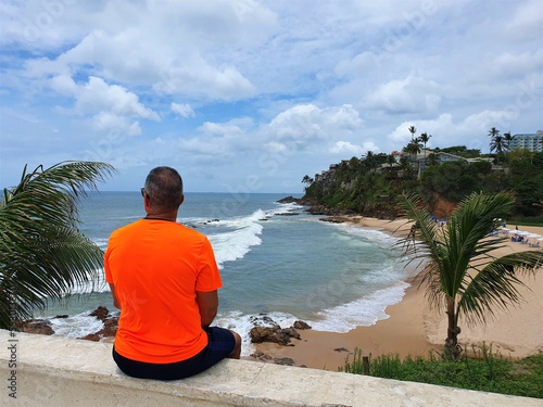 man sitting on a wall watching the beach on a blue sky day with clouds
