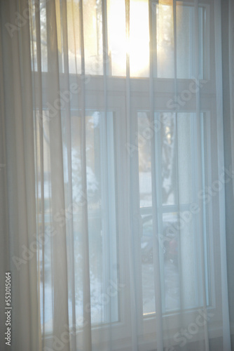 wooden window behind white silk curtains through which the orange sun of a cold winter day shines.