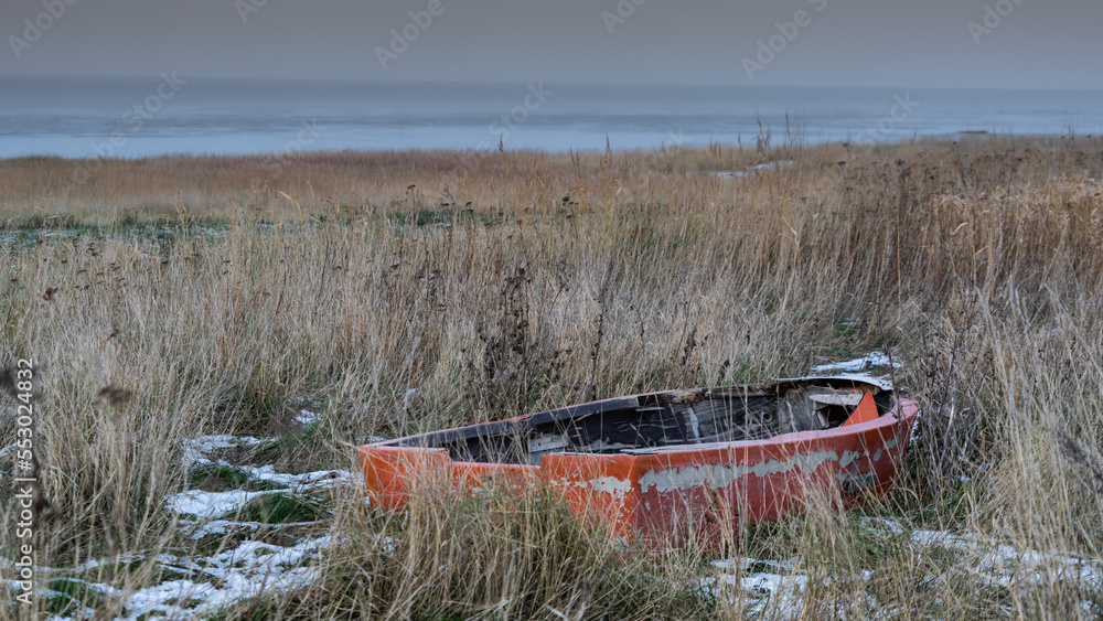 An abandoned old boat with peeling orange paint lies between clusters of beach grass and patches of snow unter a grey december sky on the coast of the Wadden Se An abandoned old boat with peeling orange paint lies between clusters of beach grass and patches of snow unter a grey december sky on the coast of the Wadden Se