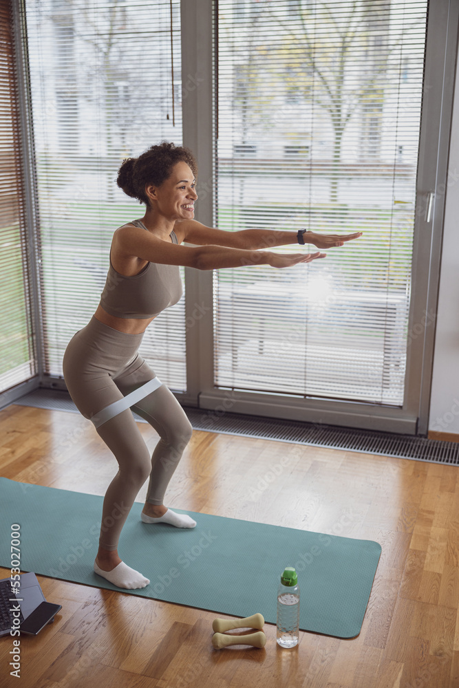 Slim woman doing exercises for legs with elastic band by online tutorials on laptop in living room Slim woman doing exercises for legs with elastic band by online tutorials on laptop in living room