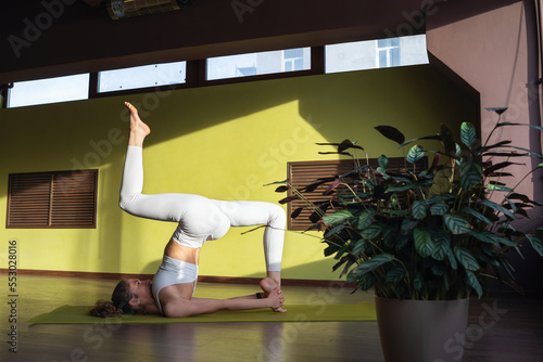 A young woman leading a healthy lifestyle and practicing yoga, performs the Ekapada Patihasana exercise, trains in white sportswear in the room on the mat photo