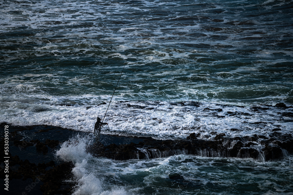 Fototapeta premium Silhouette of a man fishing in the Atlantic Ocean during high tide.