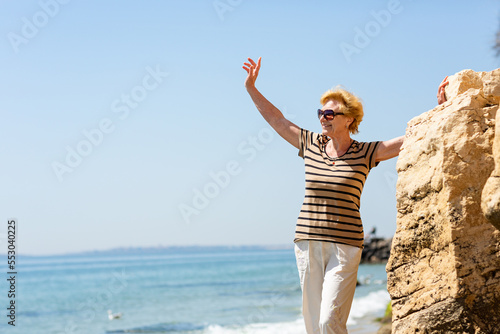 Elderly attractive smiling woman standing on a rock on the seashore waving her hand happily and enjoying life