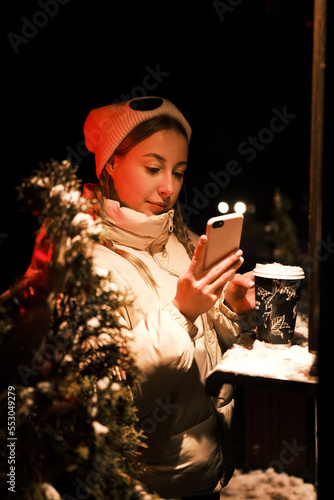 Model blonde with tails with cup of coffee near coffee shop with red and yellow light looking in smartphone. Girl weared white dress  jacket and hat posing near cafe in winter night