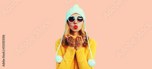 Winter portrait of beautiful woman blowing her red lips sending sweet air kiss wearing heart shaped sunglasses, yellow knitted sweater and white hat with pom pom on pink background
