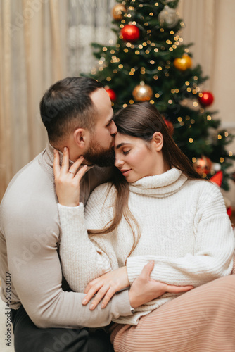 Couple in love is sitting and hugging near the Christmas tree at home at Christmas. Young man with beard is kissing head of her wife.  © VikaNorm