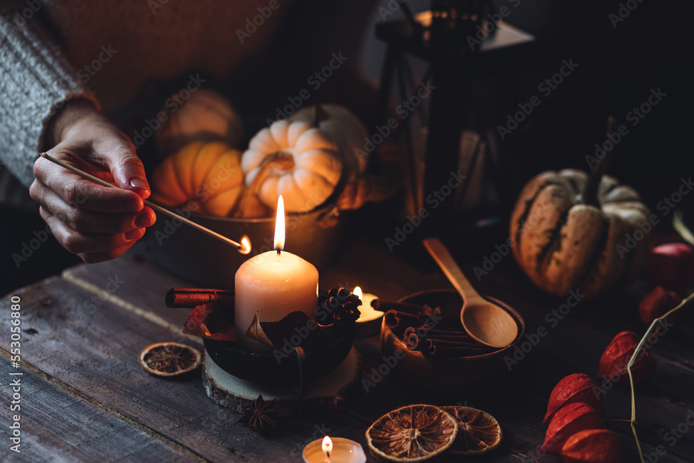 Young woman in the knitted warm sweater lights a candle on a wooden table. Cozy fall atmosphere, beautiful autumn home decor with pumpkins and leaves