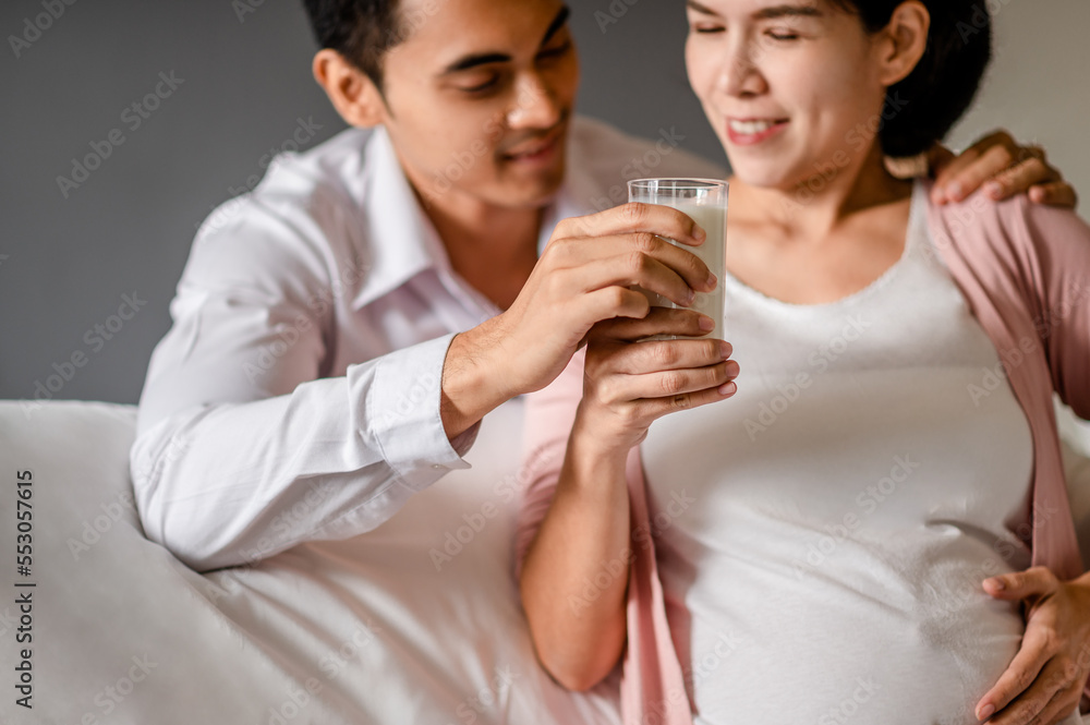 Portrait of Pregnant Asian woman sitting on a white bed Her husband holds a glass of milk and sends her fresh milk in a clear glass for her to drink. To nourish the baby in her womb. Portrait of Pregnant Asian woman sitting on a white bed Her husband holds a glass of milk and sends her fresh milk in a clear glass for her to drink. To nourish the baby in her womb.