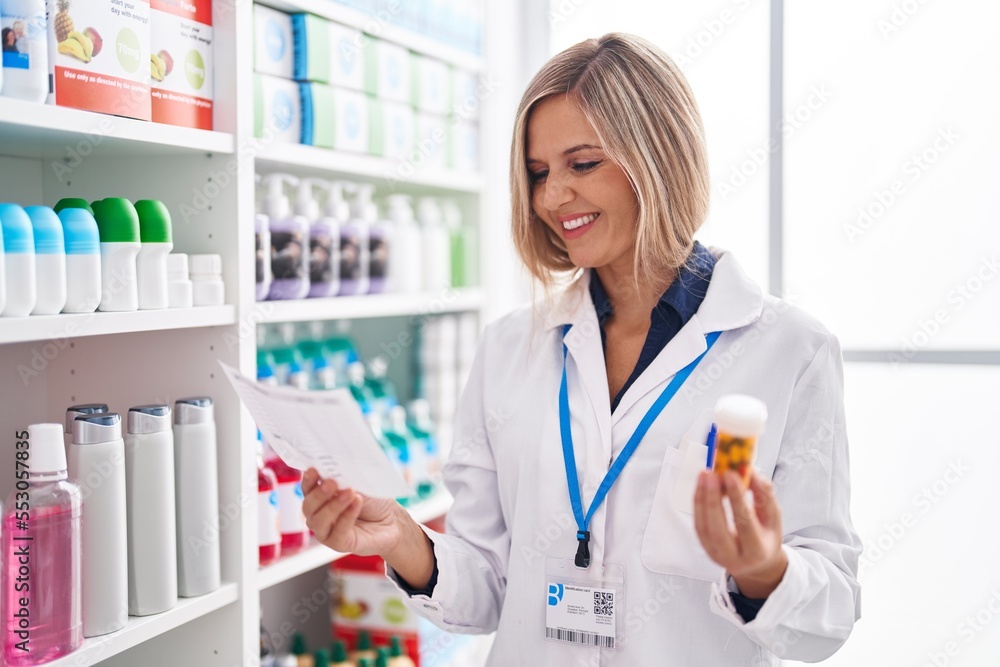 Young blonde woman pharmacist holding pills bottle reading prescription at pharmacy