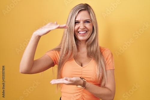 Young woman standing over yellow background gesturing with hands showing big and large size sign, measure symbol. smiling looking at the camera. measuring concept.