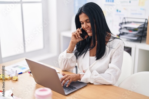 Middle age hispanic woman business worker using laptop talking on smartphone at office