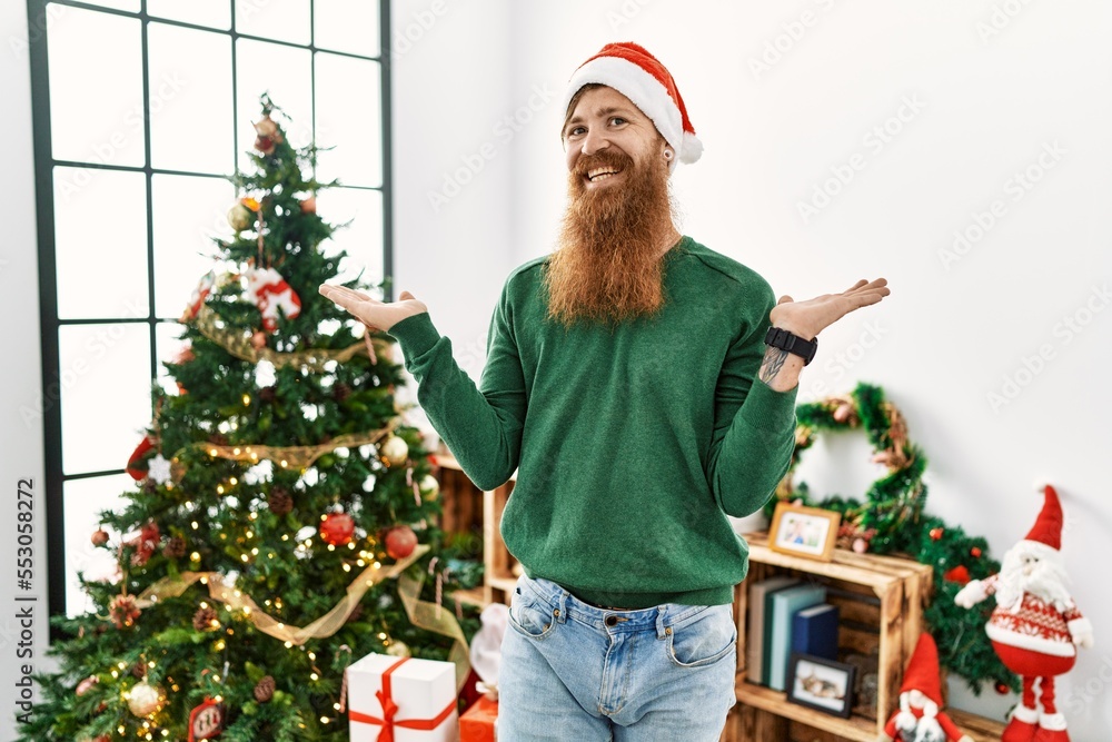 Redhead man with long beard wearing christmas hat by christmas tree smiling showing both hands open palms, presenting and advertising comparison and balance Redhead man with long beard wearing christmas hat by christmas tree smiling showing both hands open palms, presenting and advertising comparison and balance