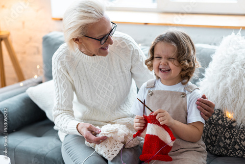 Grandmother and granddaughter doing craft toys and knitting near decorated Christmas new year tree. Cute little girl and attractive senior woman at home in the living room on the sofa having fun