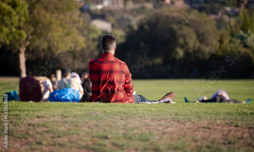 A man wearing a red flannel shirt sitting on grass at a park