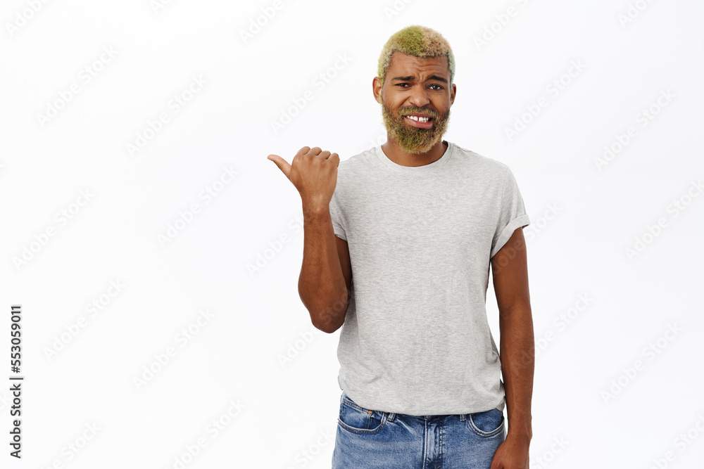 Naklejka premium Dislike. Disappointed african american guy with yellow modern hairstyle and beard, pointing left, grimacing from disappointment, white background