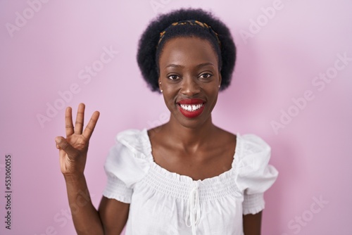African woman with curly hair standing over pink background showing and pointing up with fingers number three while smiling confident and happy.