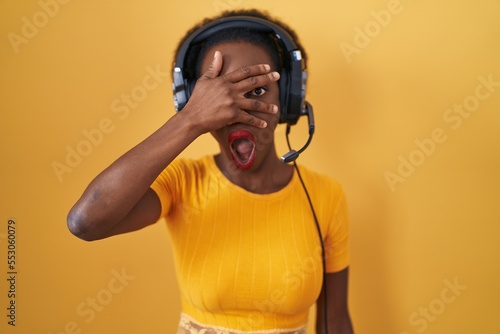 African woman with curly hair standing over yellow background wearing headphones peeking in shock covering face and eyes with hand, looking through fingers with embarrassed expression.