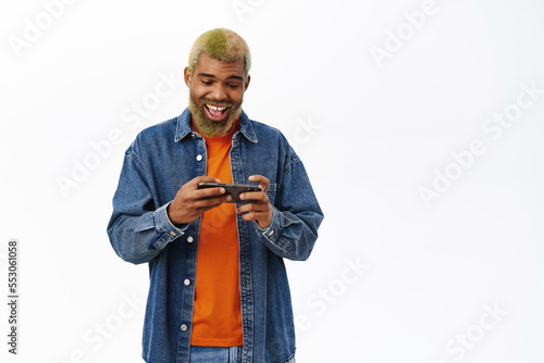 Hipster guy plays on mobile phone, using smartphone with excited face, white background