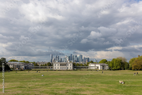 Canary Wharf view from Greenwich Park, filled with people enjoying leisure activity on a cloudy summer day, wide shot.