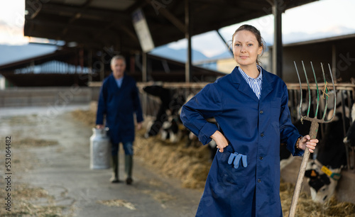 Portrait of confident female livestock farm worker in blue work coat posing with pitchfork in hands 