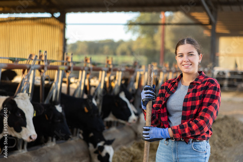 Portrait of positive young woman farmer standing in cowshed, holding handle of working tool and smiling.