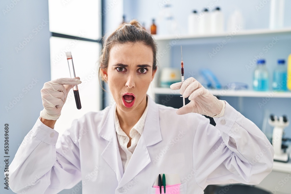 Young woman working at scientist laboratory holding blood sample in shock face, looking skeptical and sarcastic, surprised with open mouth Young woman working at scientist laboratory holding blood sample in shock face, looking skeptical and sarcastic, surprised with open mouth